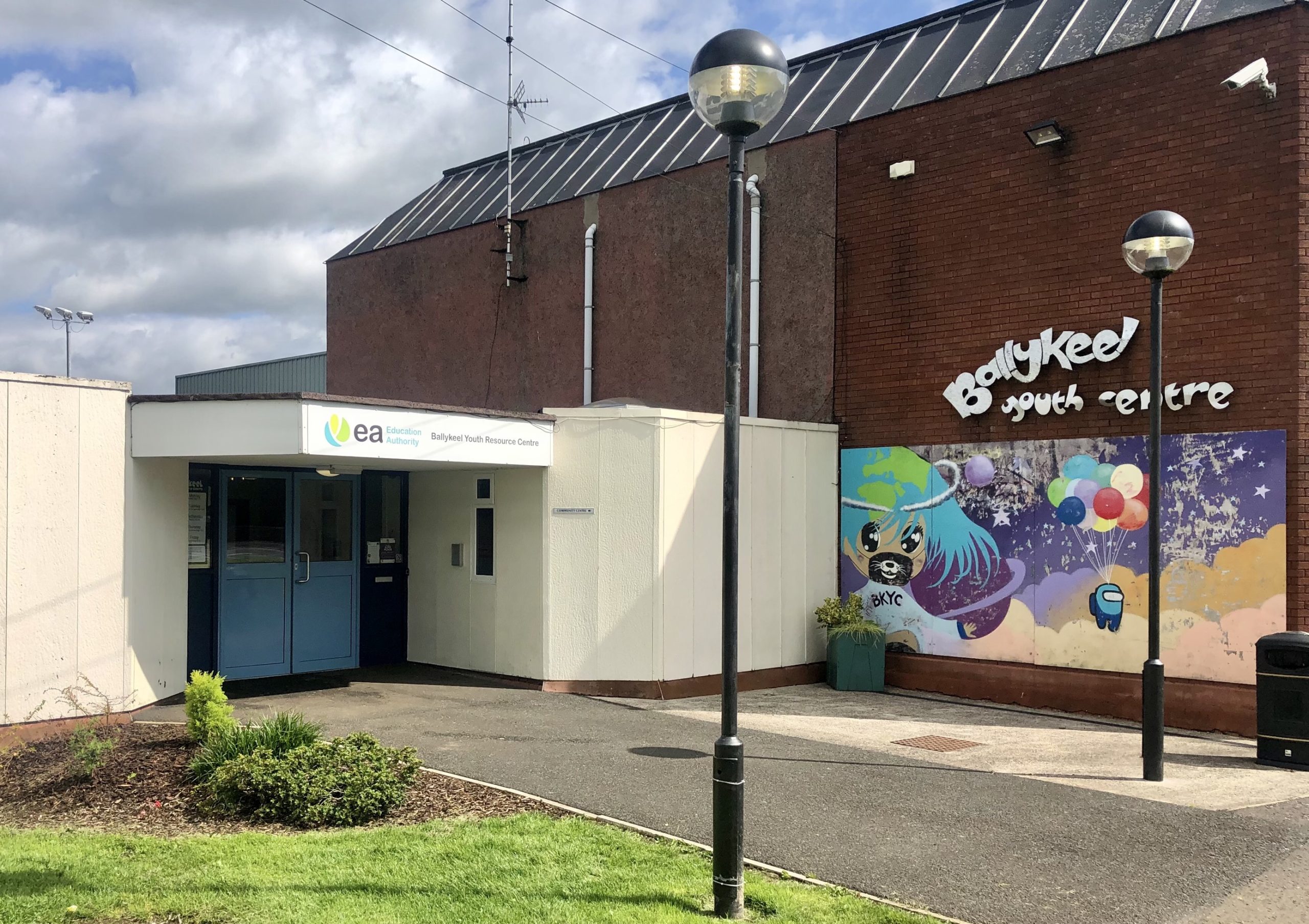 Front of Ballykeel. The door is on the left side and there is a large mural of a bear and a robot with balloons. There is a large "Ballykeel Youth Centre" sign above the mural