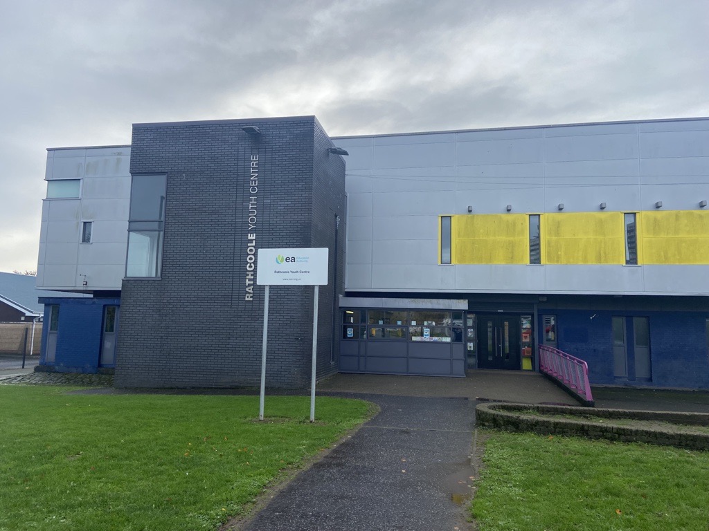 Front of Rathcoole. A large grey building with some yellow panelling on the front. There is grass in front of the building.