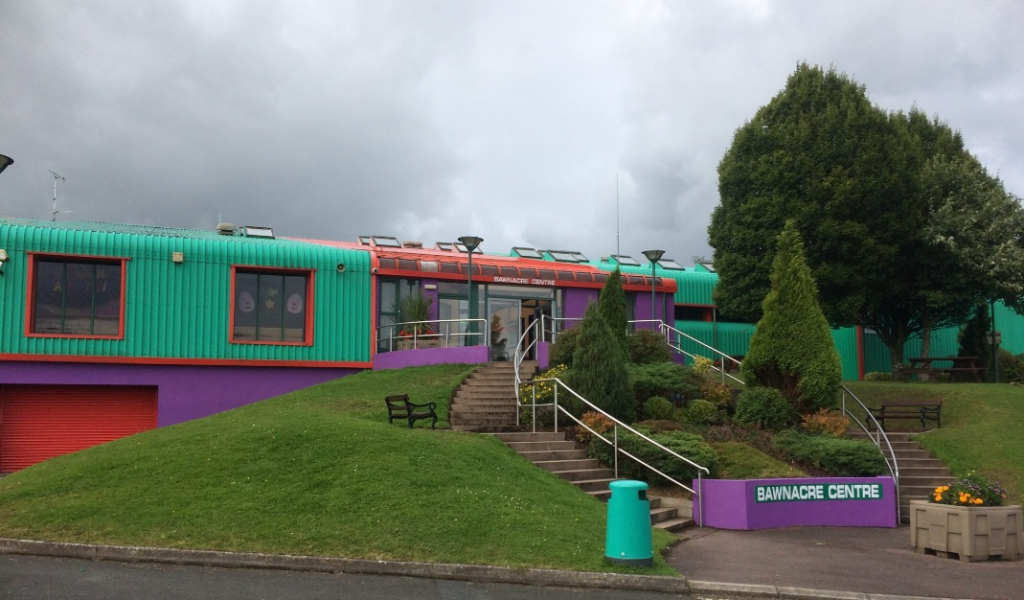 Front of Irvinestown centre. There are steps up to the front door with grass and plants on either side. The building is green, purple and red.
