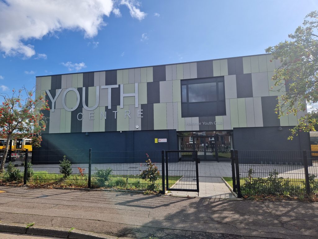 Front of Hammer Youth Centre. The building has large green, grey and cream tiles across the front with a large "Youth Centre" sign. There is some grass and plants to the front.