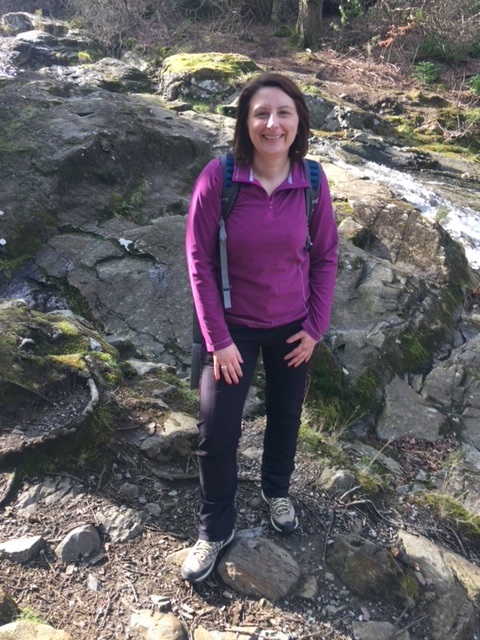 Joanne standing on a rock while hiking. She is wearing a purple jumper and rucksack